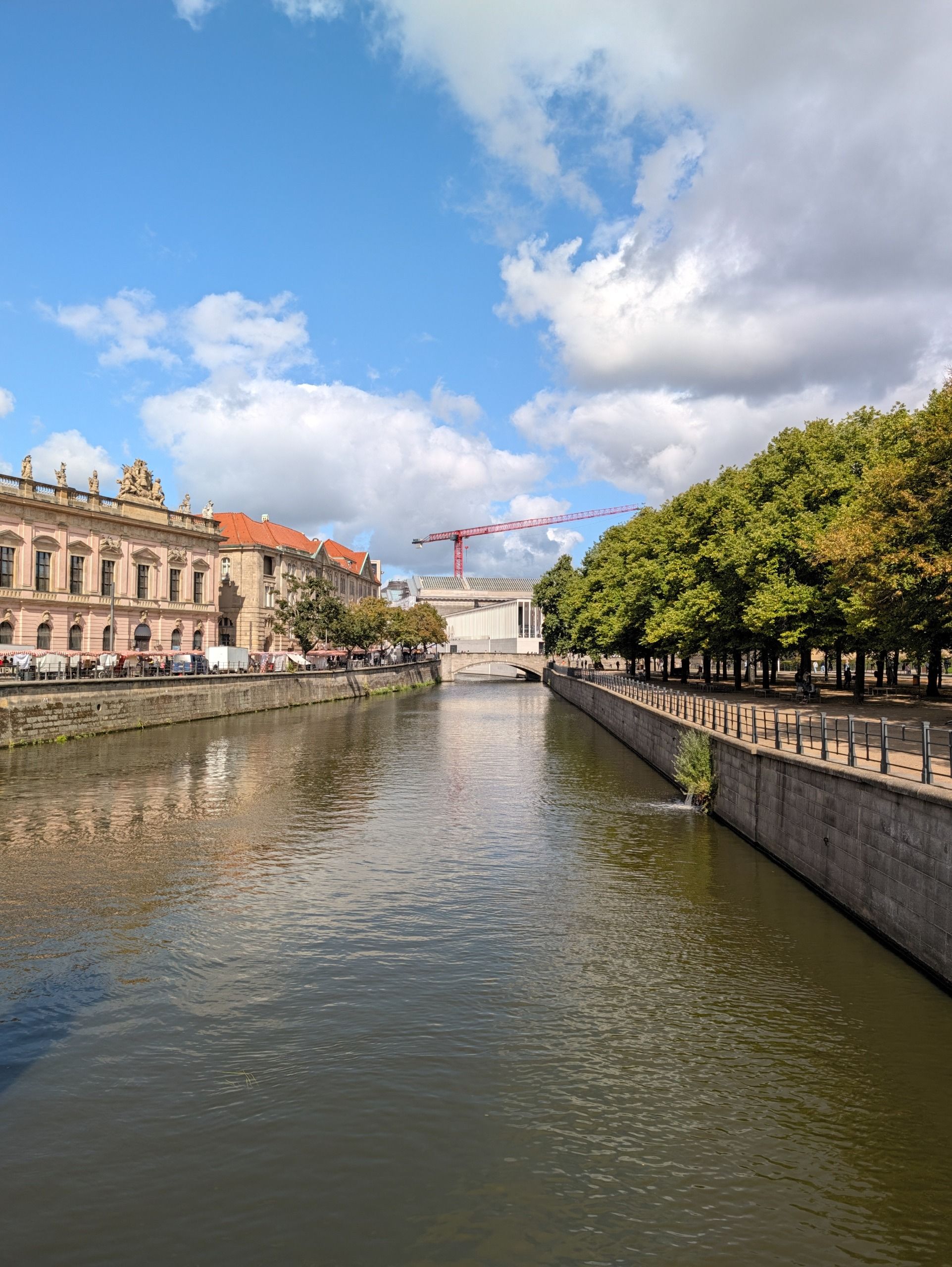 Canal in Berlin lined with historic buildings and trees beneath a bright blue sky with large clouds.