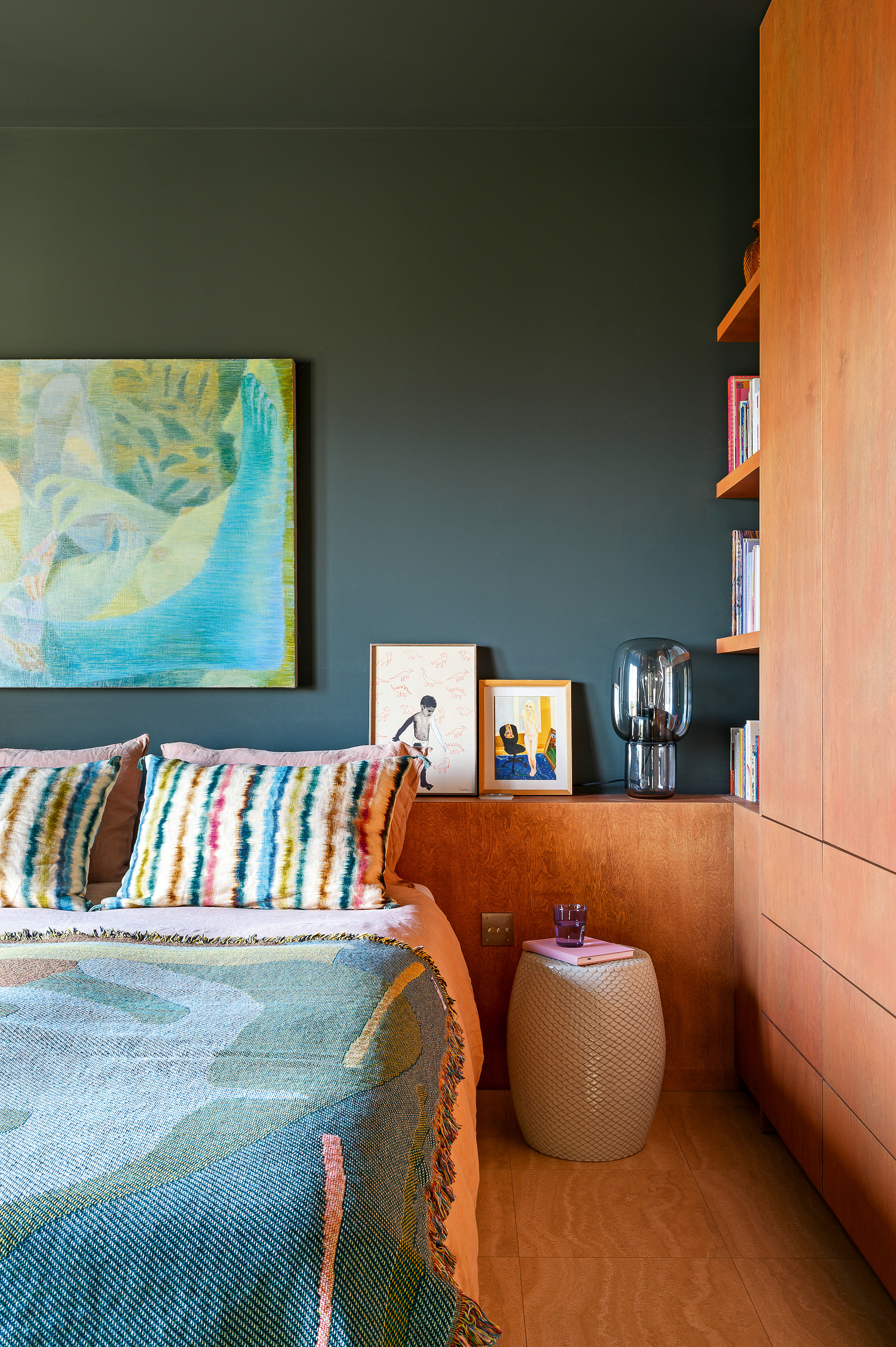 Bedroom with cherry wood built-in cupboards and headboard, dark teal walls, turquoise and green artwork and green and teal throw