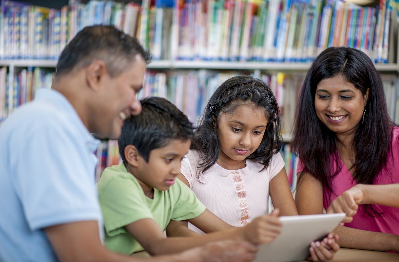 A mother and father are helping their son and daughter with their homeschooling assignments at the library. They are watching an educational video on a digital tablet together.