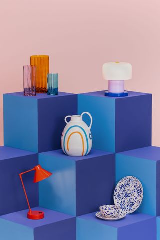 A selection of decor items on a blue stand, including a red angle-poise lamp, blue speckled stoneware plates, and colourful glassware