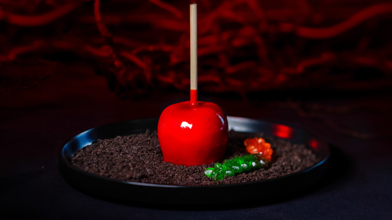 La manzana venenosa mostrada en un plato, sobre una mesa negra, frente a una cortina roja para Universal Halloween Horror Nights 33.