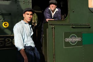 Kieran Fish (Dane Williams) stands leaning against the cab of a green steam train, while Harry Bishfield (Richard Glover) is inside the lab leaning out. Both are dressed in shirts, overalls and flatcaps, although Kieran's overalls are unrolled down to his waist.