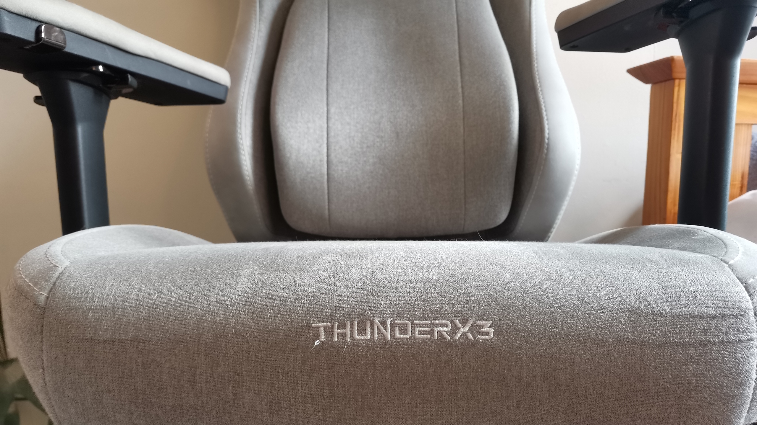 The ThundercoreX3 Core gaming chair in grey in front of a pale wall, next to a wooden bed frame