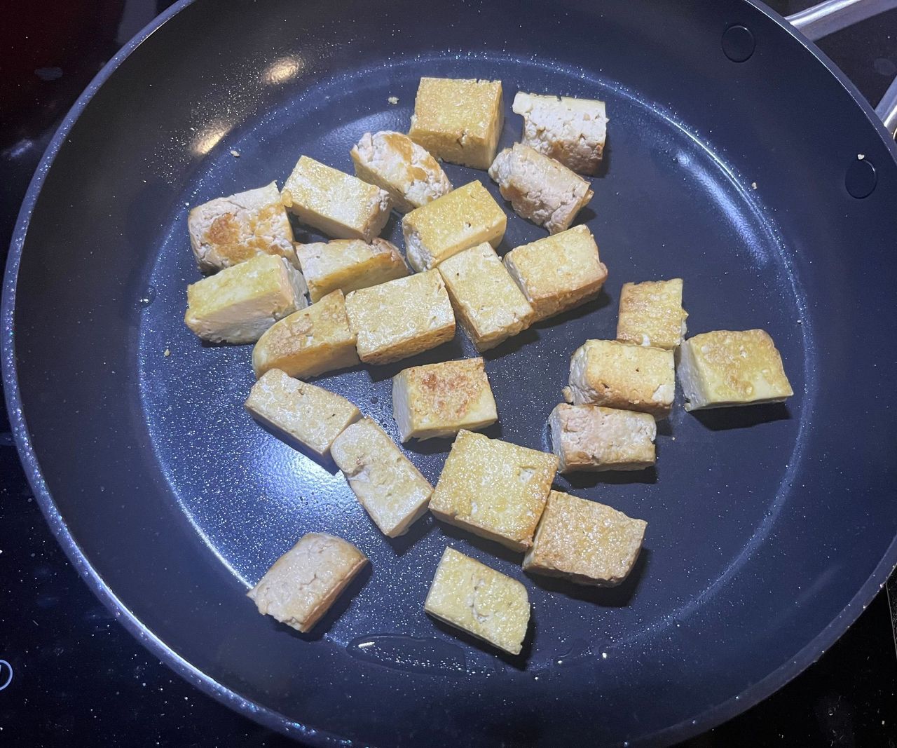 A black ceramic frying pan on a black stovetop with cubes of tofu frying inside.