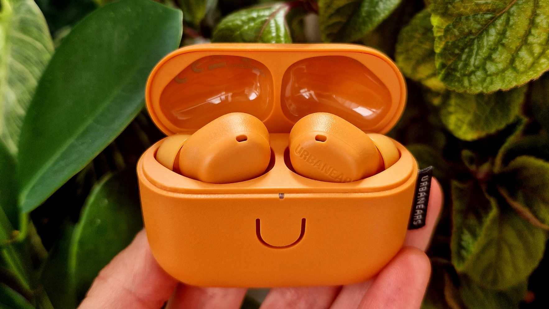En hand håller upp ett par orange Urbanears Juno som ligger i sitt tillhörande laddningsfodral framför ett gäng gröna växter.