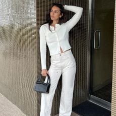 A woman wearing white jeans, a white cardigan, and black handbag.