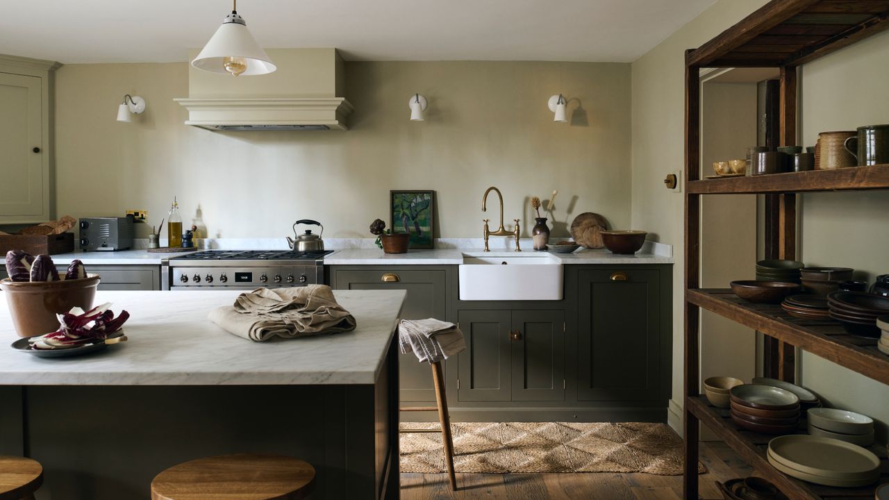 dark green kitchen cabinets and pale green walls, wooden shelving unit with tableware, wooden floorboards, marble countertop, butler sink, brass faucet, white wall lights, island, stools, rug