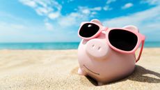 A piggy bank wearing sunglasses sits on the beach.