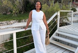 A woman wearing a white linen vest and pant set with a raffia handbag and gold hoops.