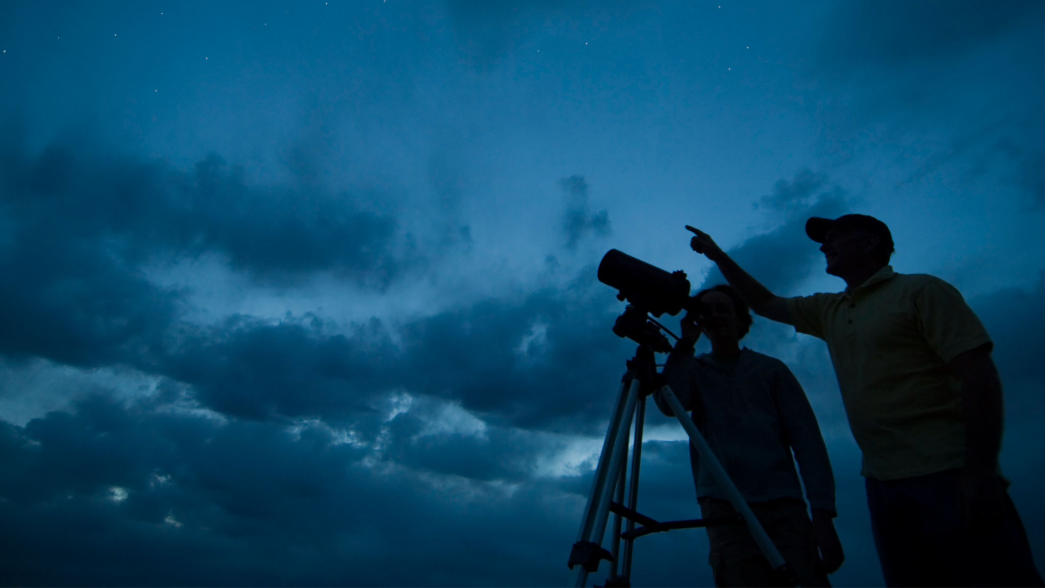 A couple silhouetted against a twilight sky using a telescope.