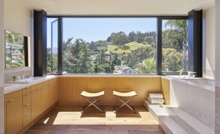 A bathroom in a residential home with wide windows that gives a view of other homes and the hills . The bathroom has an oak laminate and and concreate design. The concrete bath features 2 stairs