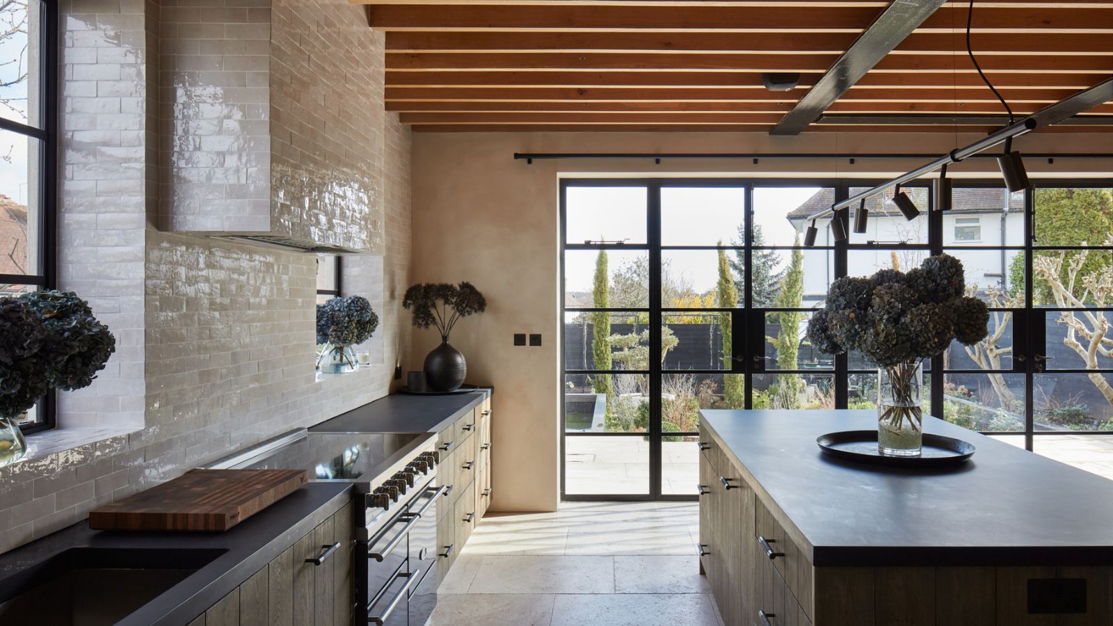 Light-filled kitchen with timber ceiling beams, pale tiled walls and a large island. Black-framed glass doors open onto a garden beyond.