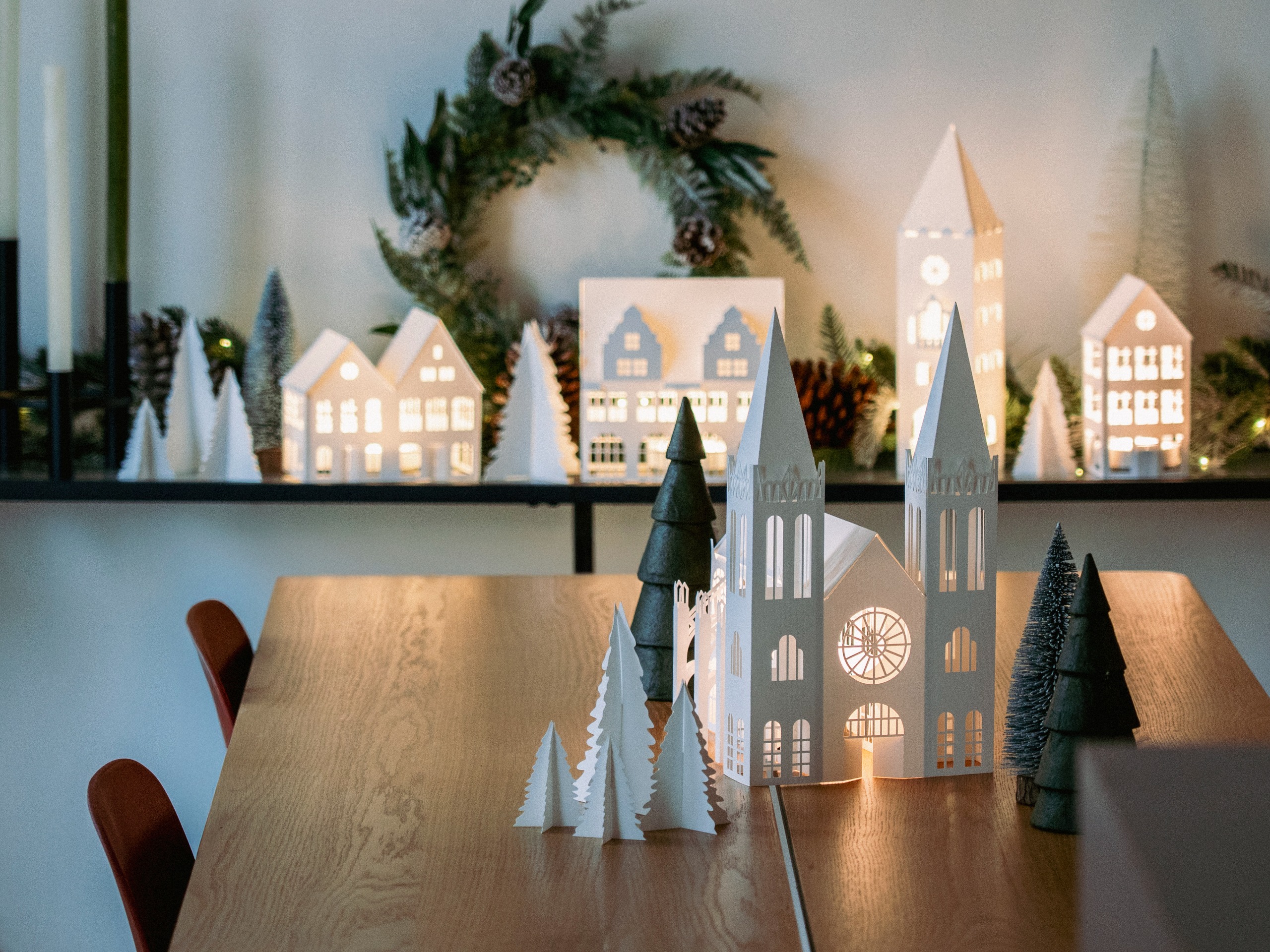 A christmas village of lit-up white paper decorations, some of which are on a wall shelf in front of a wreath among taper candles and other decorations, while the rest are on the table in among trees