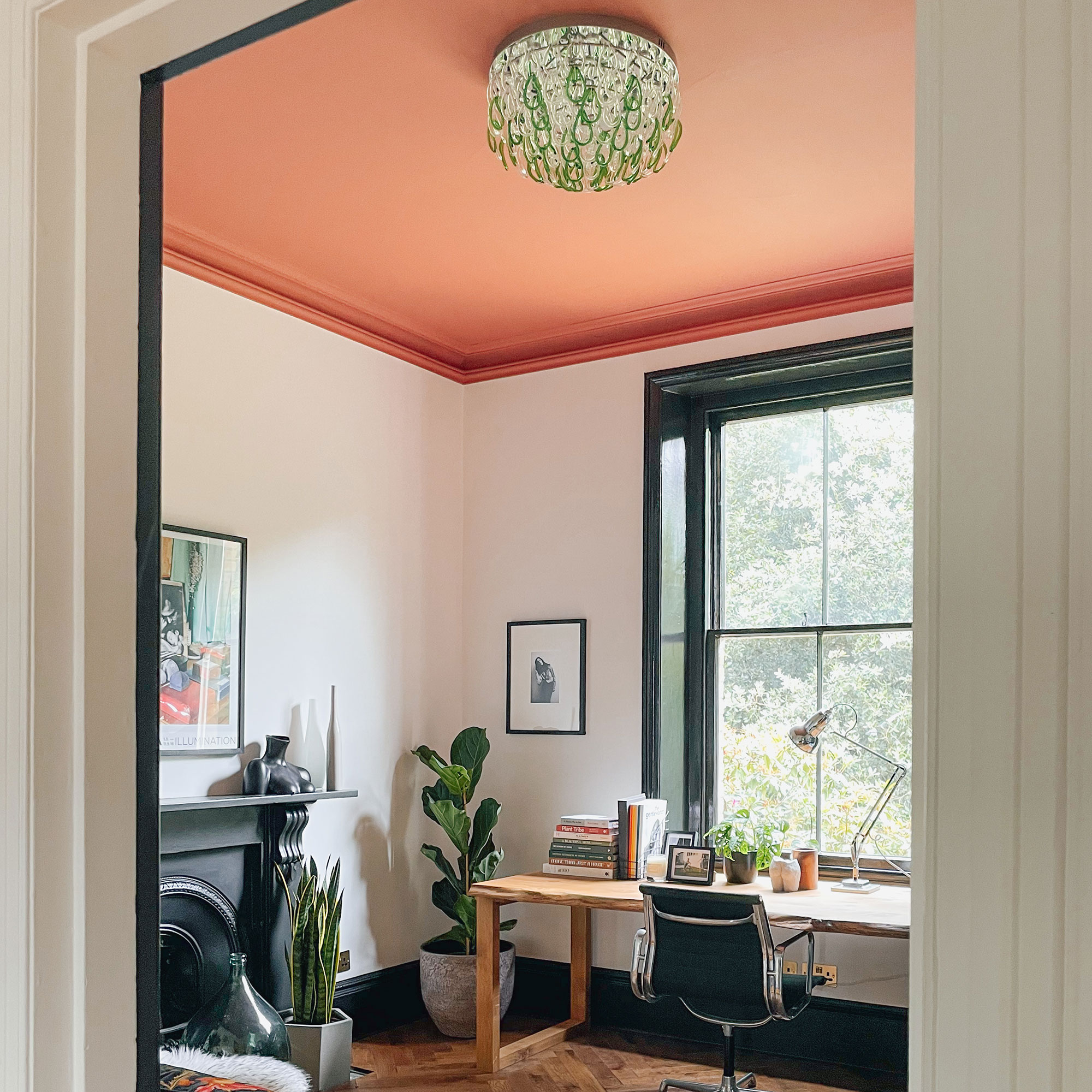Home office with green and clear glass pendant light, orange-red ceiling, neutral walls, houseplants, black painted fireplace and wooden desk in front of large window