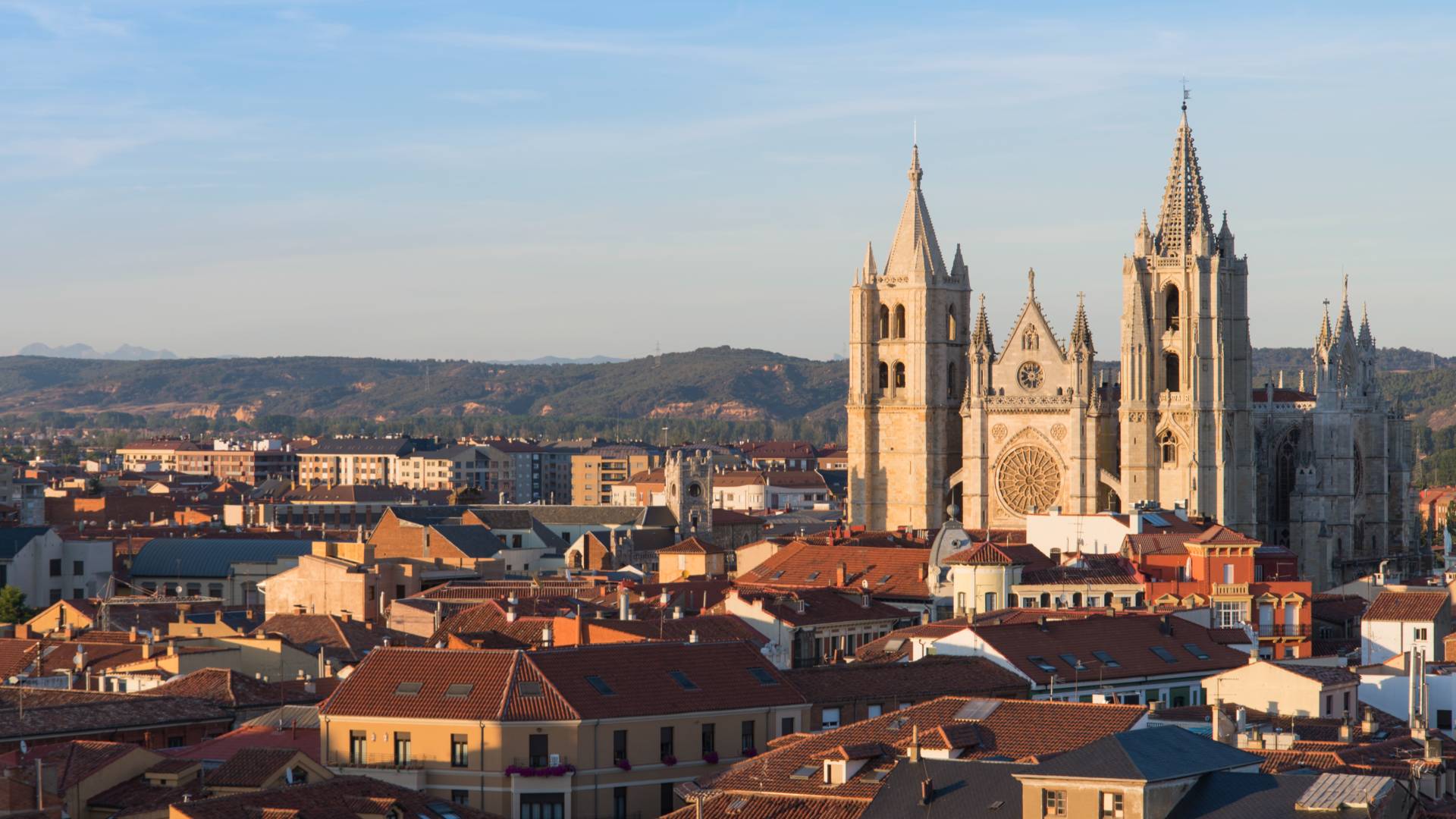 A panoramic view of a city with red-roofed buildings and the Le&oacute;n Cathedral, a Gothic church with tall spires and ornate details, set against distant hills under a clear blue sky.