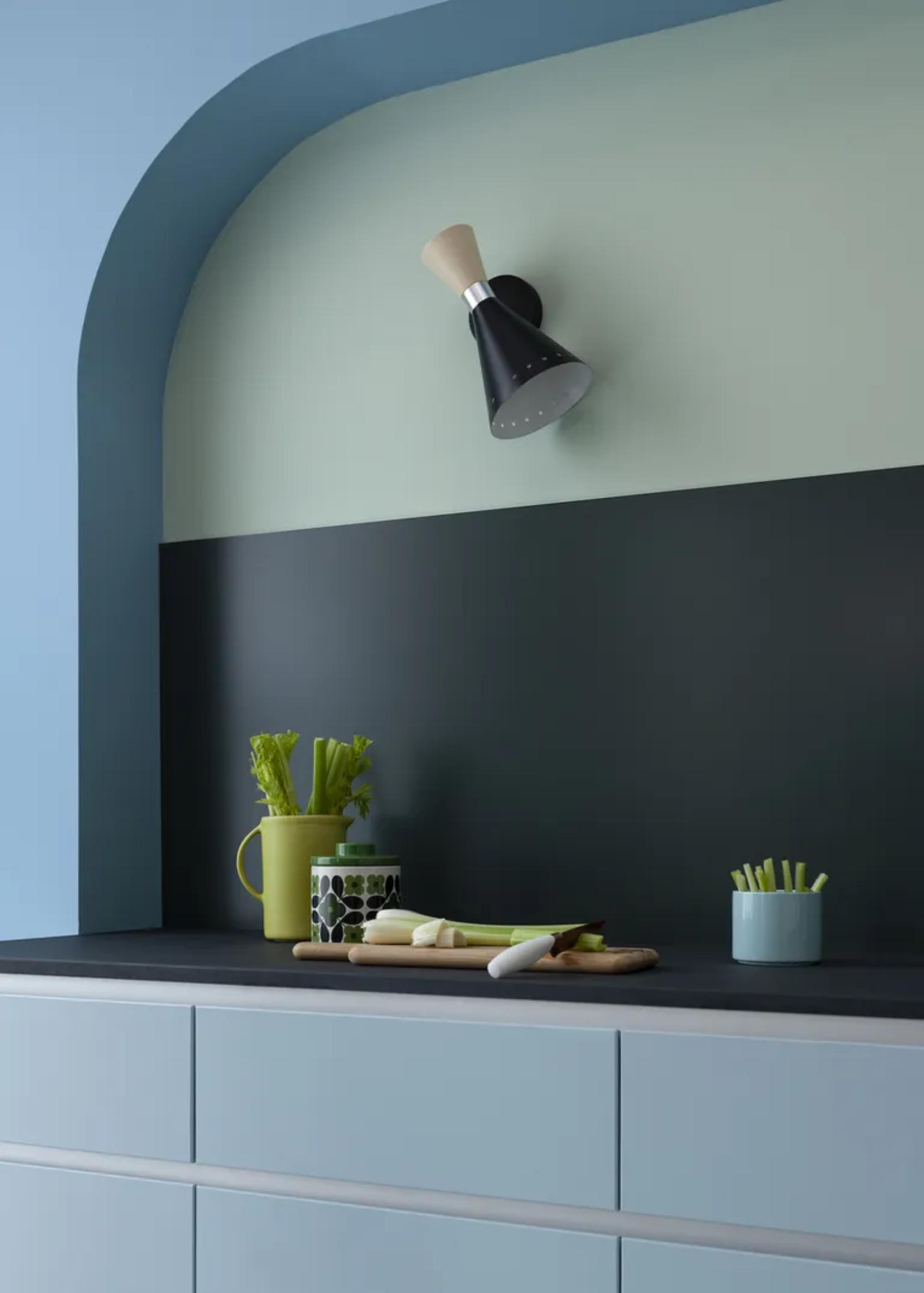 A kitchen with a sky blue wall, pale blue drawers, a dark gray backsplash and counter, and a pale teal color block with a black wall sconce. A cutting board with a white knife, chopped leeks, a yellow jug of leeks, a patterned jar, and a bowl of greens are also seen.