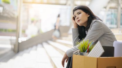A businesswoman sits on the steps of a building with a box of belongings next to her.