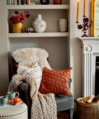 A living room with a gray armchair with a beige throw and orange pillow on top of it and a beige wall behind it with vases and books on the shelves