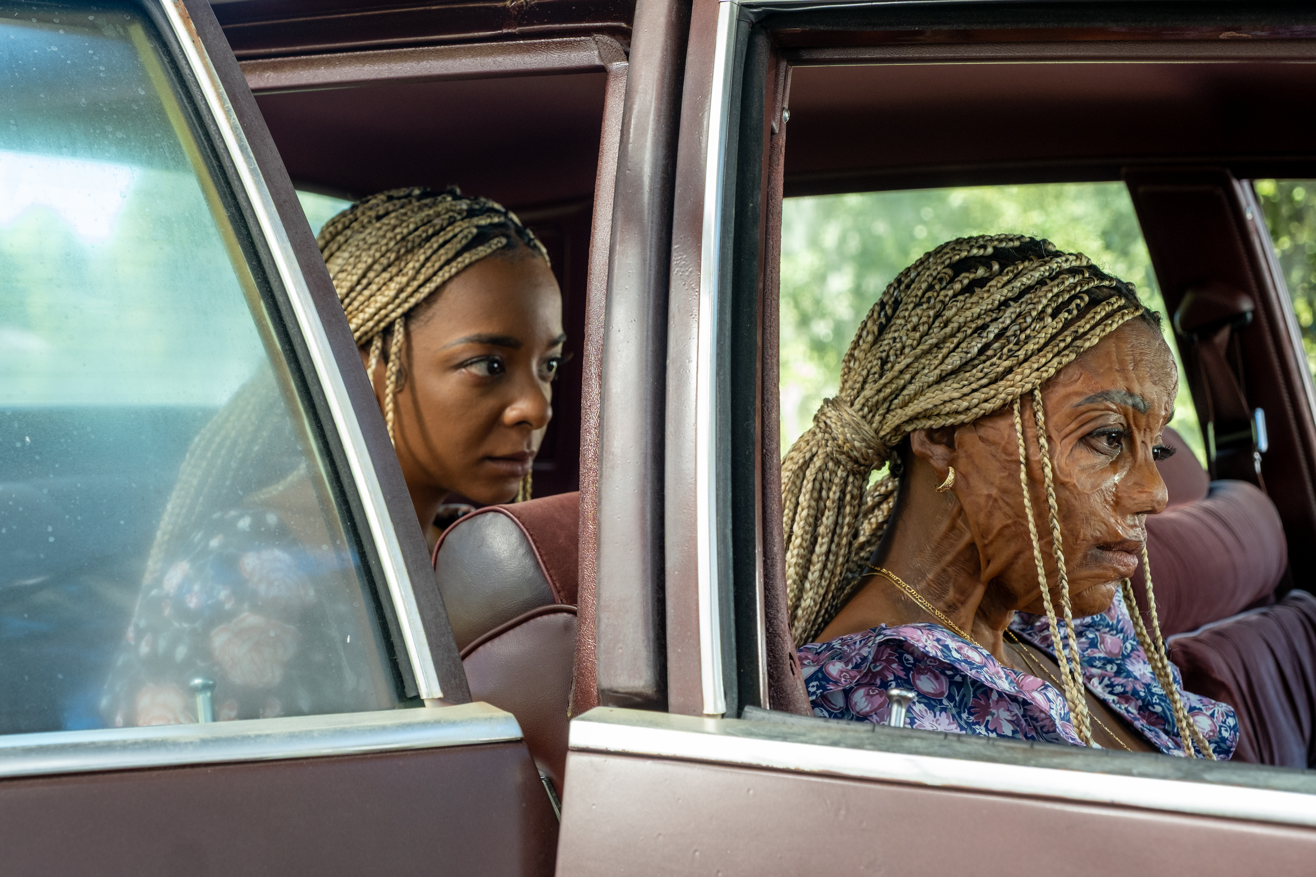 A Black woman with blonde box braids (Kara Young as Racine) sits in the back seat of a car. Another woman with facial scars (Mallori Johnson as Anaia) sits in the front seat. A still from 'Is God Is.'