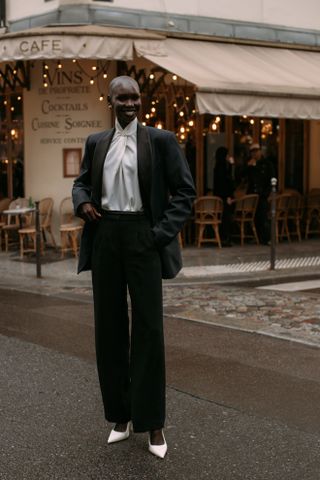 A woman standing in the street in Paris wearing a black suite, white blouse, and white heels