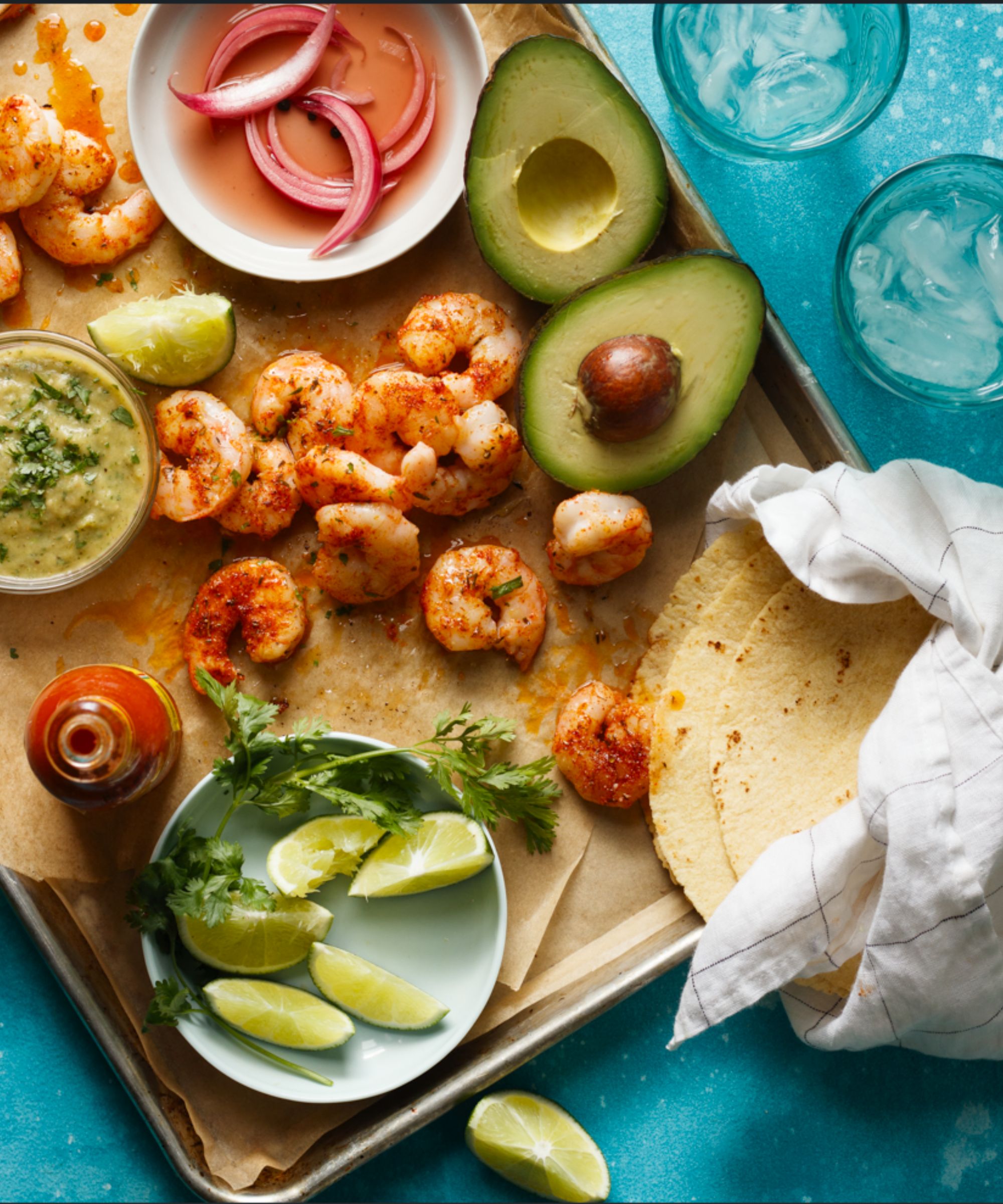 Tray with shrimp, lime wedges, avocados, tortillas and sauce with two glasses of ice water beside it