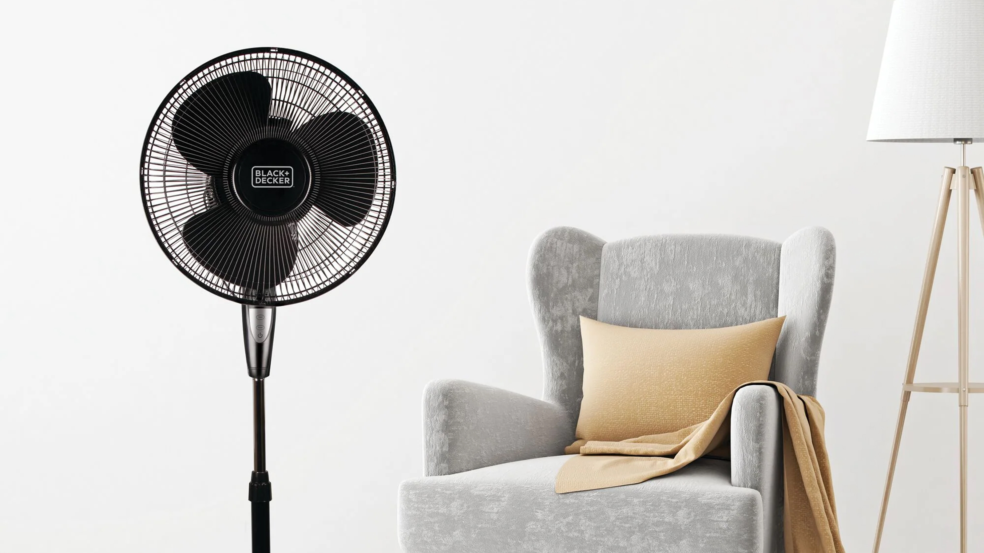 A black BLACK+DECKER pedestal fan in a white room with a grey armchair and floor lamp