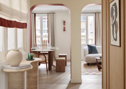 A hallway with neutral walls, console table and Roman blinds with a deep red fringed trim, leading into a living room with curtains lined with deep red edge