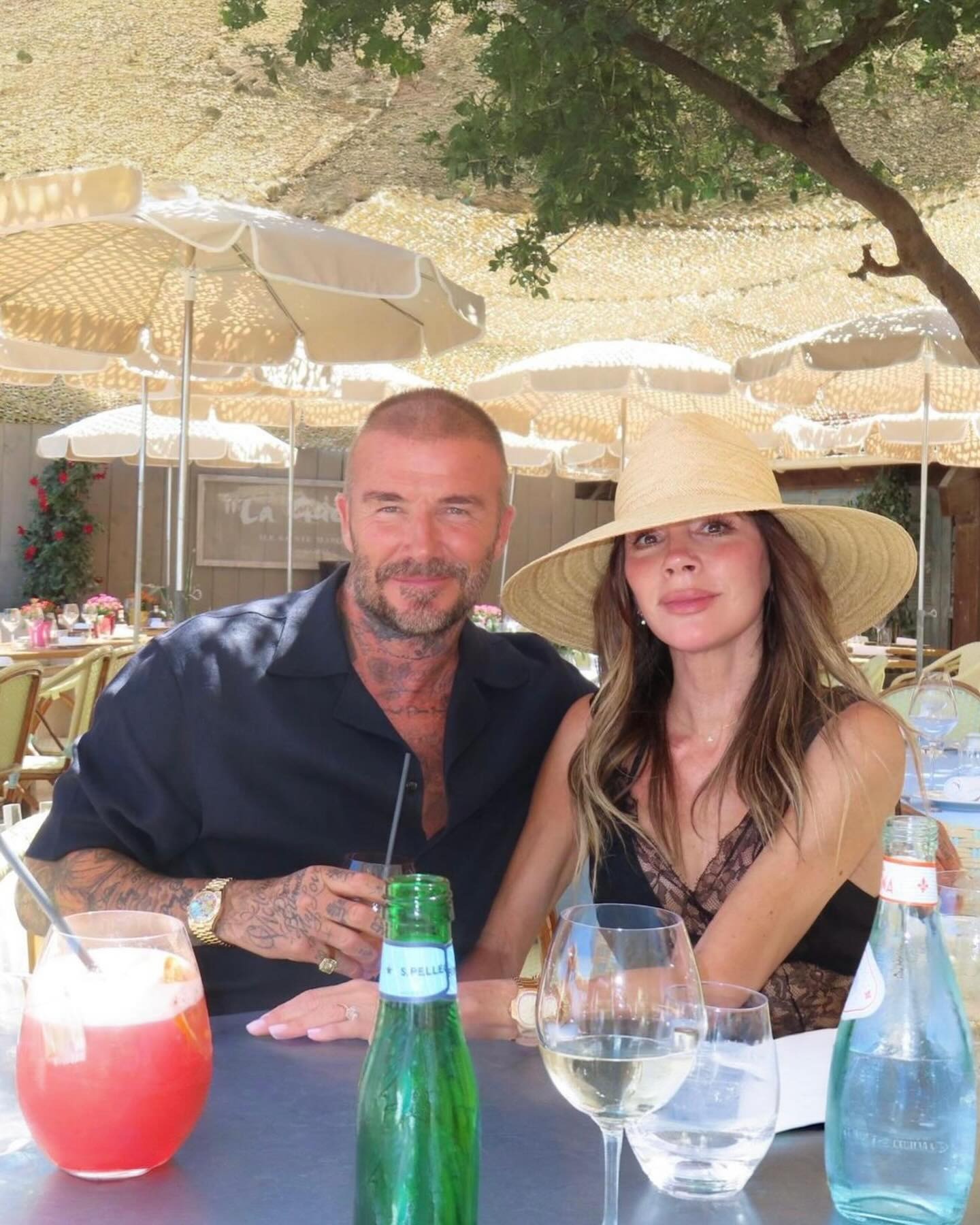 Victoria Beckham sits next to David Beckham wearing a lace trim black dress with an oversized straw hat.