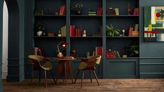 Image of a library room painted in a dark blue paint color. There are built-in bookshelves lining the walls and a table and two chair set.