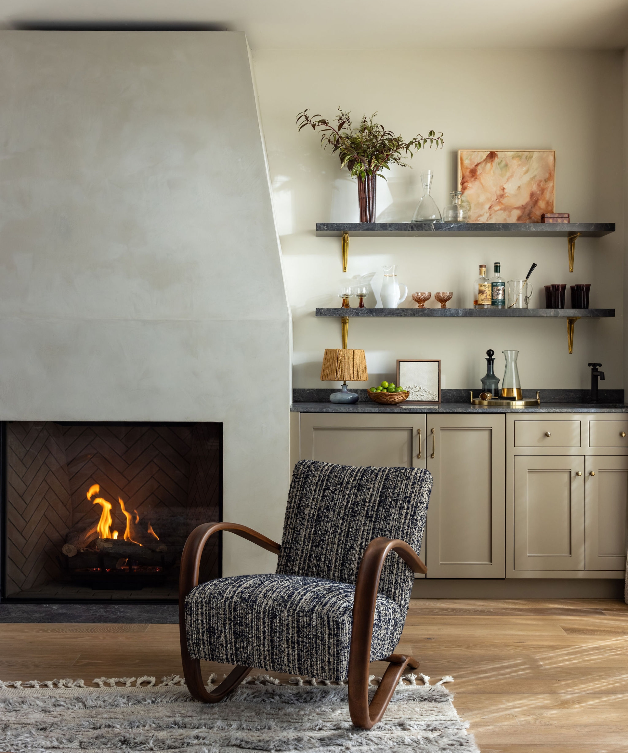 Living room with a curved wood armchair in blue patterned fabric, a plaster fireplace and a built-in bar with stone shelves.
