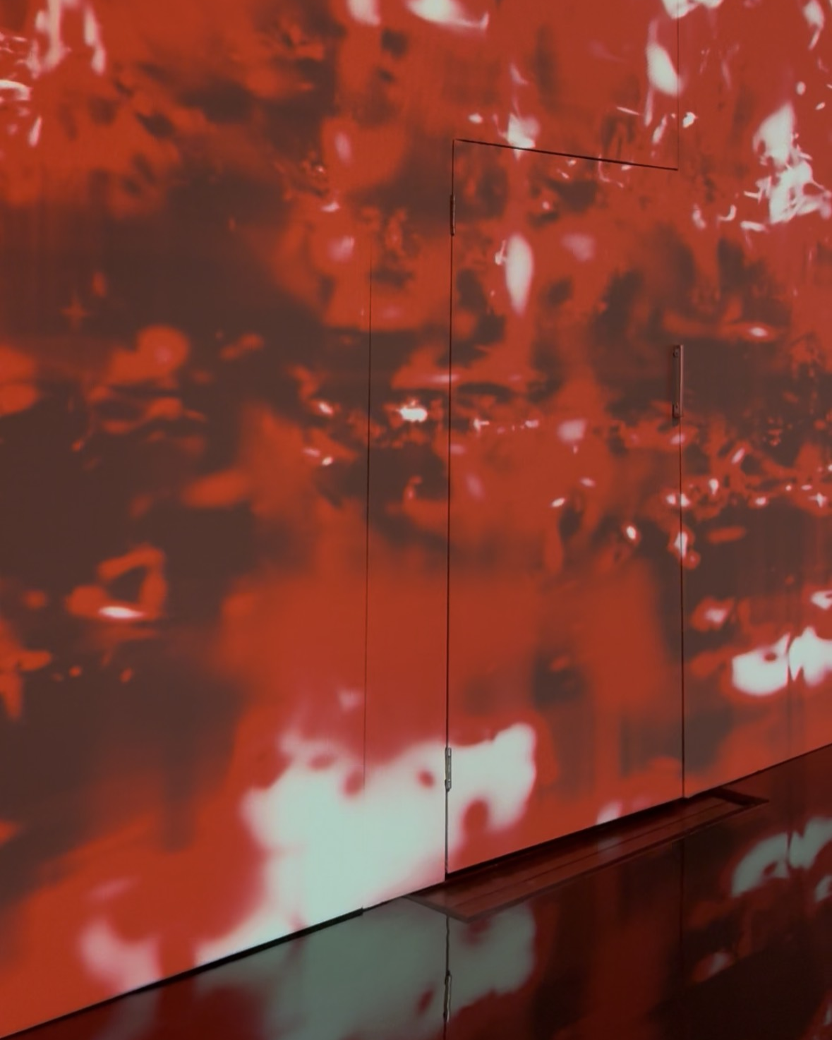 An audiovisual installation projects abstract forms in red, black, and white onto a plain wall.