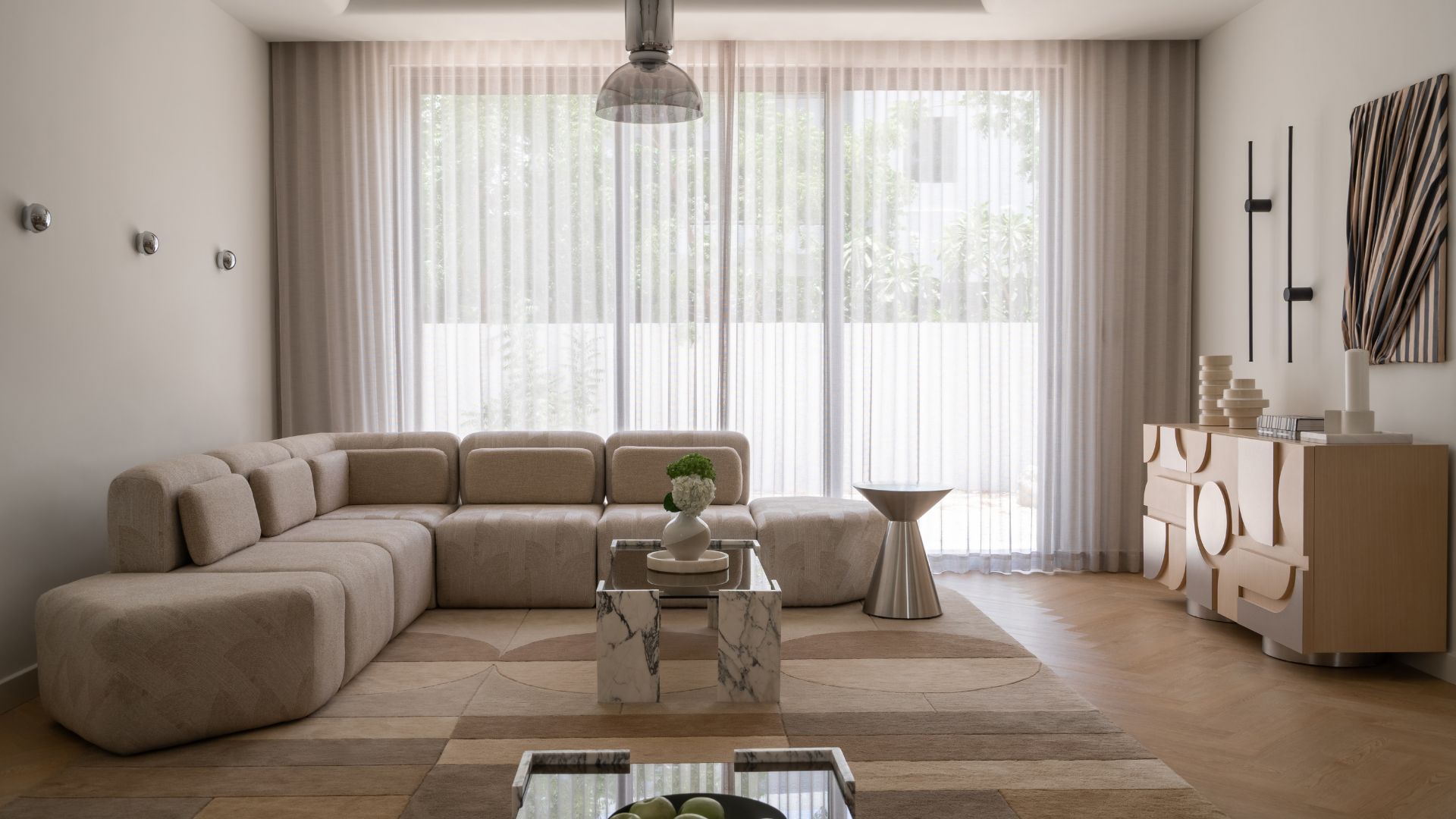 A minimalist living room with a rounded beige sectional a marble and glass coffee table, a steel side table, a wood and silver sideboard, a set of three chrome sconces, and a geometric rug