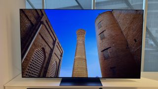 The LG G4 OLED TV photographed on a white stand in a showroom, with a soundbar positioned in front. On the screen is the blue ceiling of a building. On the screen is a tower between two large buildings.