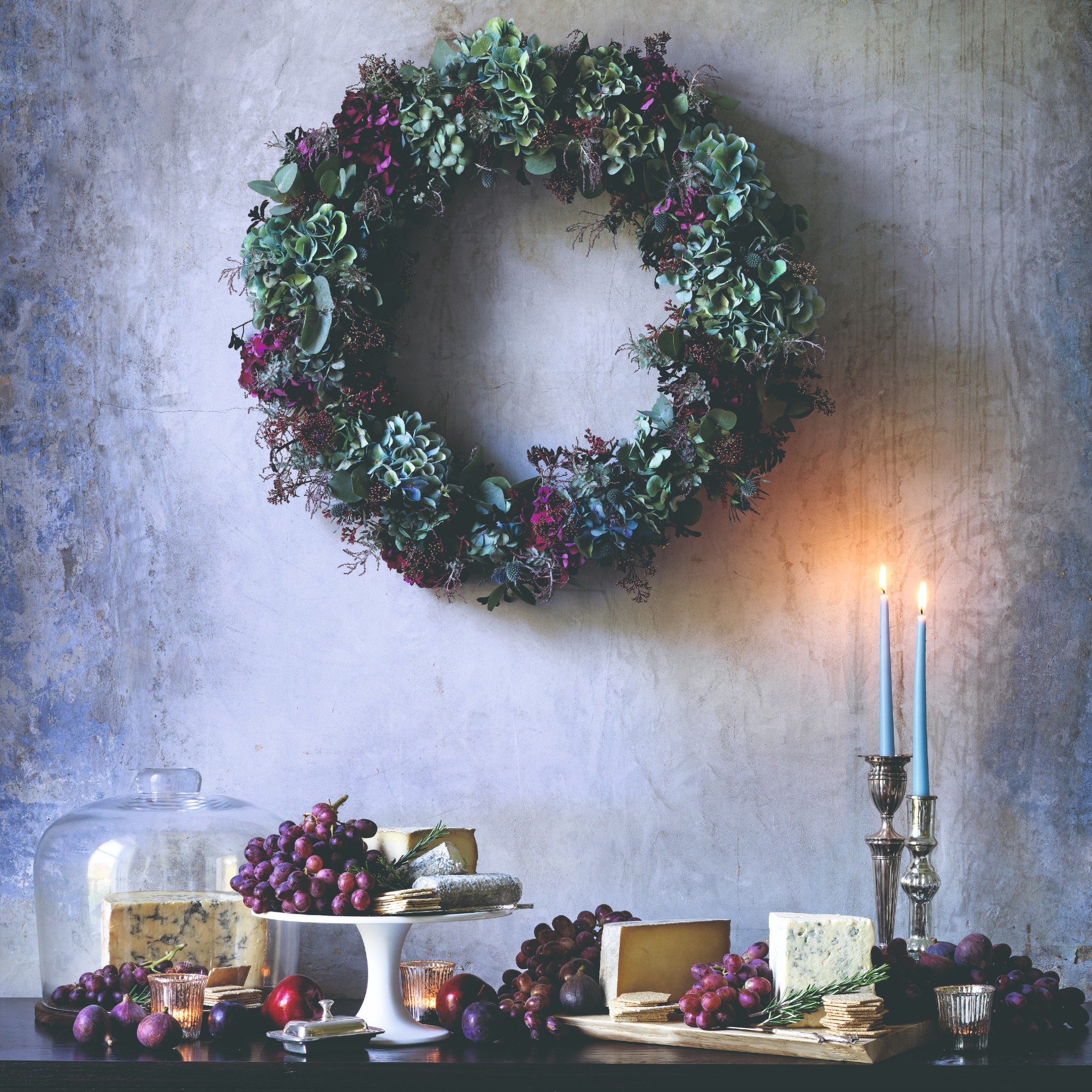 A dining table decorated with grapes and cheese and candles and a Christmas wreath hung on the wall above