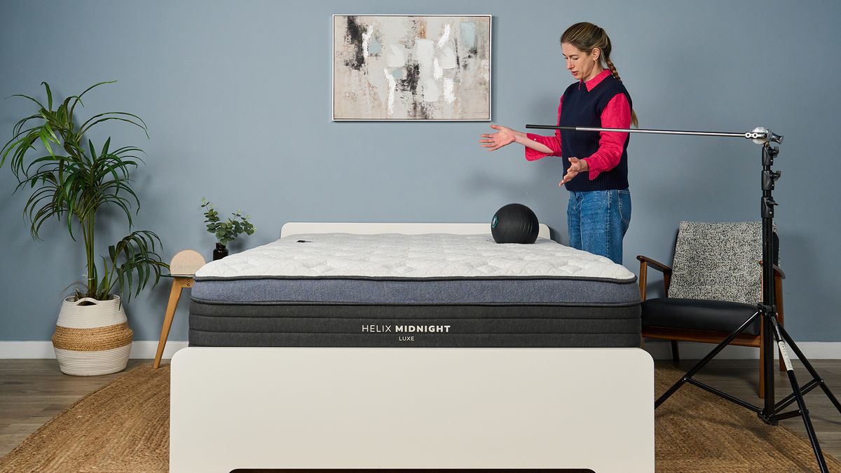 Tester Nicola Appleton drops a 5kg slam ball on the Helix Midnight Luxe mattress to measure motion isolation. A phone with an accelerometer is taped to the other side of the mattress while a rod arm ensures she drops from the same height each time.