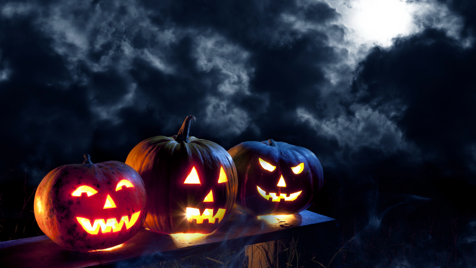 A trio of carved Jacko lanterns shine on a bench with a moonlit cloudy sky forming a dramatic backdrop.