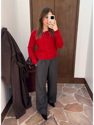 Nikki wears a red cardigan, gray trousers, and black heels.