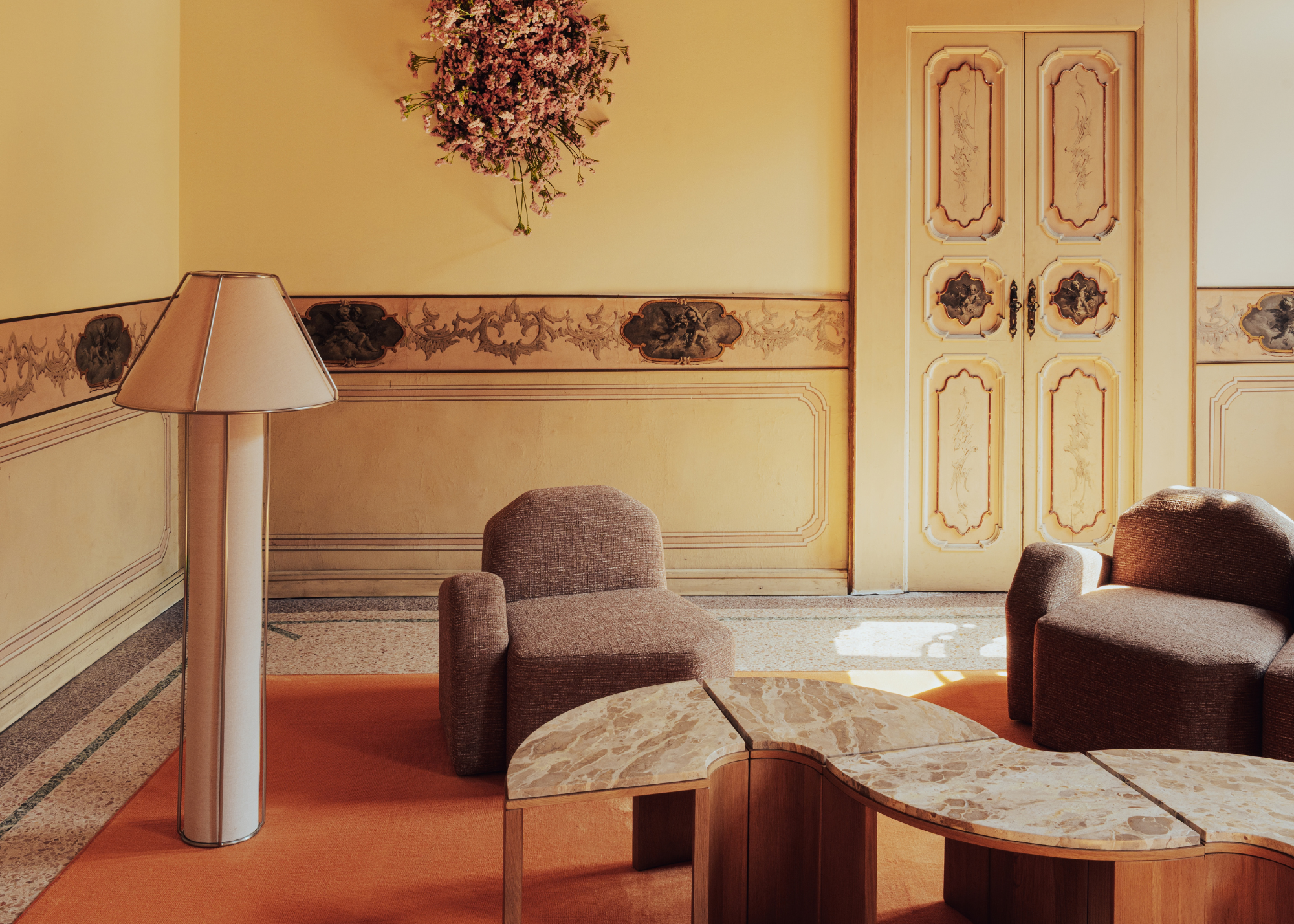 A bright butter yellow living room with traditional wallpaper accents, an ornate door, brown armchairs, a curved modular coffee table, and a metal floor lamp with a fresh bouquet on the wall