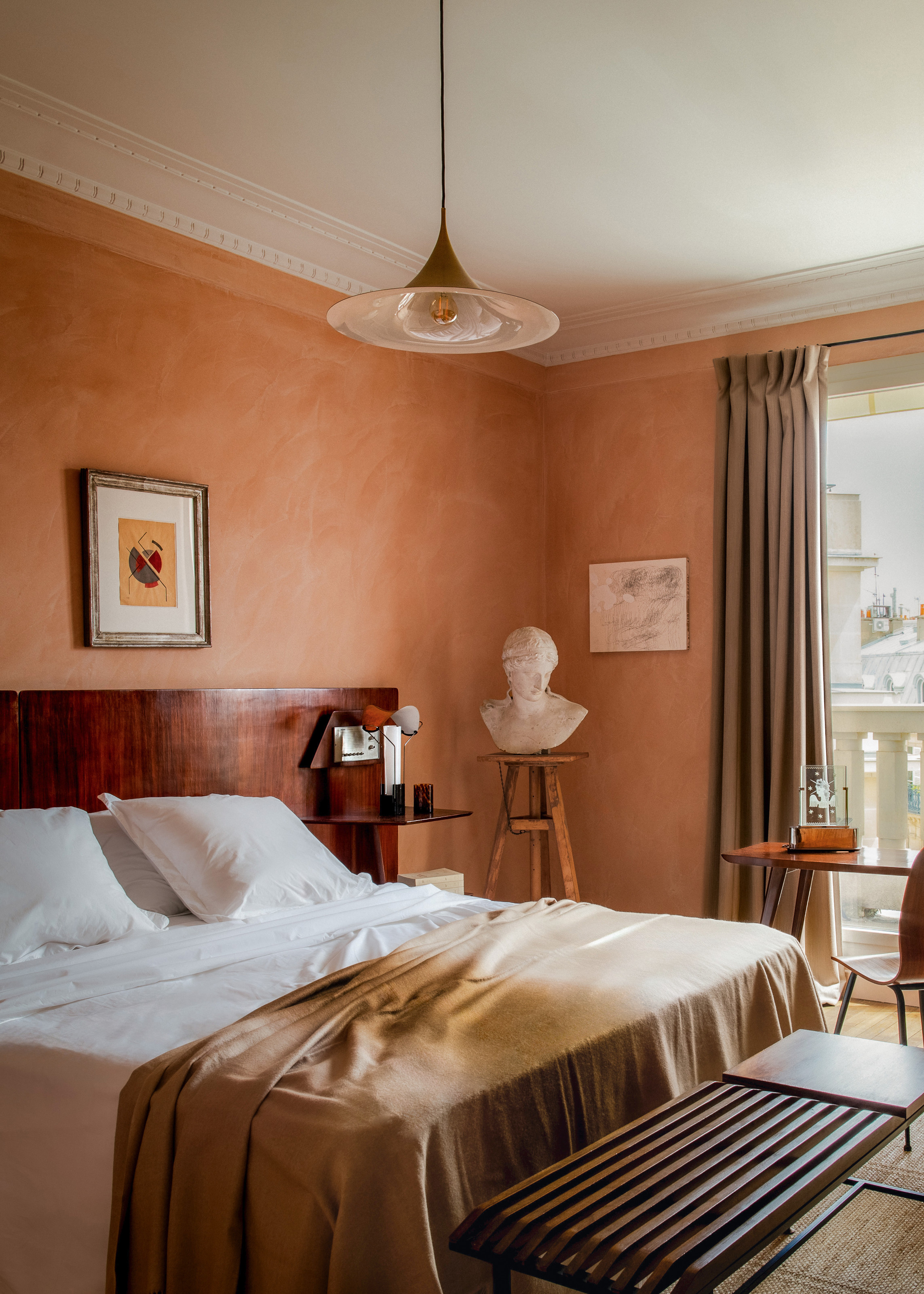 Bedroom with textured peach walls, dark walnut headboard, soft ochre throw, wooden bench at end of bed, and plaster bust on a wooden plinth in the corner