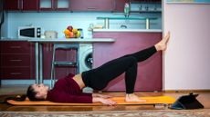 A woman performs a single leg glute bridge at home on an exercise mat. She is on her back, with her right leg bent and right foot planted on the floor, while the rest of her body is elevated in a straight line, left foot pointing upwards in a 45° angle. Behind her we see a kitchen counter, fruit bowl and microwave.