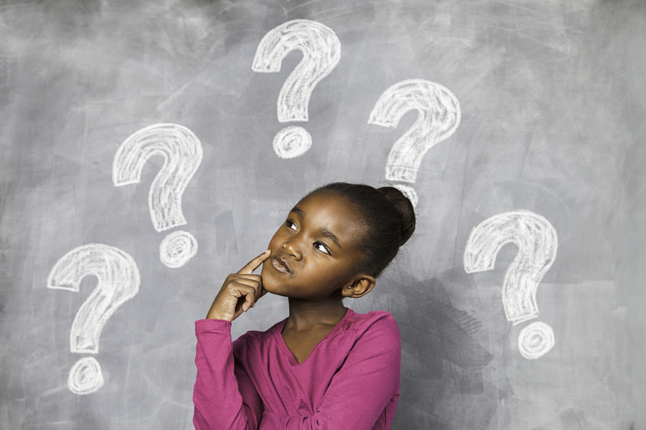 A young girl is sitting in front of a black chalkboard with question marks drawn on it. She is thinking very hard. Cape Town, Western Cape, South Africa