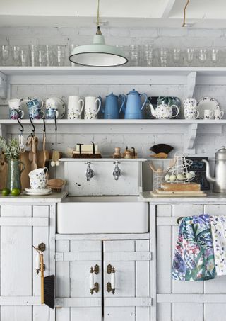 White rustic cupboards in a free standing kitchen with freestanding open shelving, full of glasses, mugs, jugs and kettles