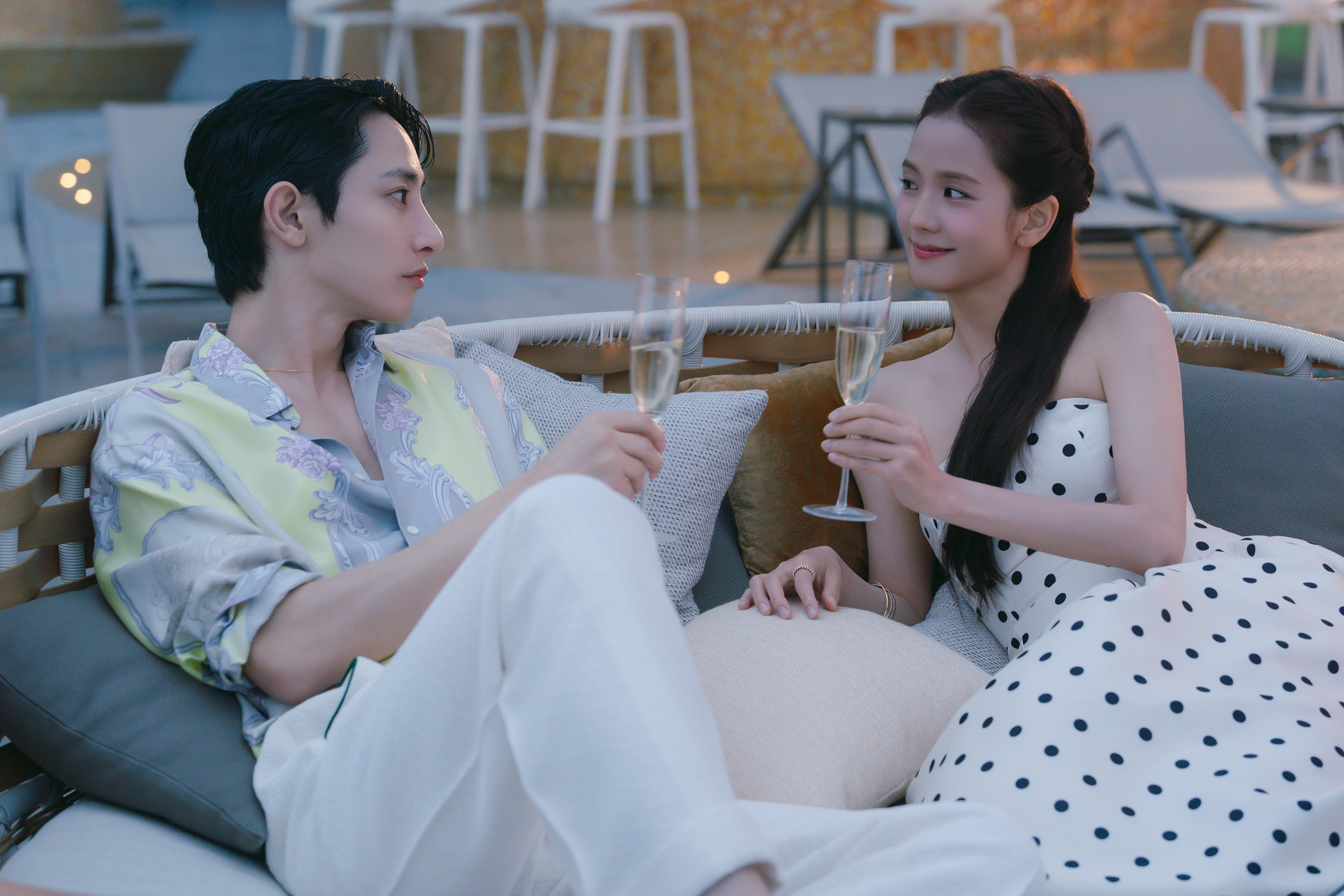 A man in a pastel shirt (Lee Soo-hyuk) and a woman in a polka-dot dress (Jisoo) toast champagne flutes while sitting in an outdoor loveseat, in the Netflix K-drama 'Boyfriend on Demand.'