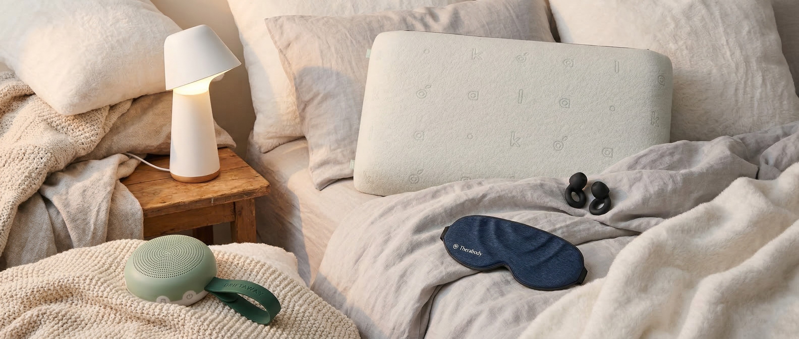 A sleep mask, ear plugs, white noise machine and pillows on a bed with a smart lamp on the beside table