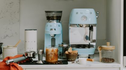 Image of Smeg's retro-style cold brew coffee machine in light blue on a white kitchen countertop. The matching grinder is to the left of it, and there is also the Smeg milk frother. There are different coffee-making accessories also on the counter.