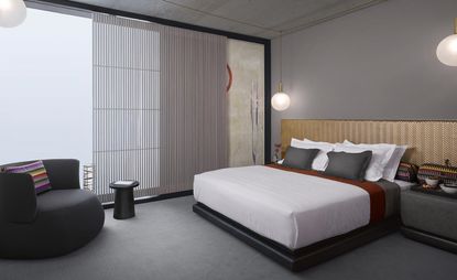 A room in the Nobu Hotel Shoreditch. The room is decorated in various shades of gray - walls, carpet, and bedside tables are in lighter gray, while the armchair and a small coffee table are in darker gray. The headboard is painted gold. Floor-to-ceiling windows cover the far wall.