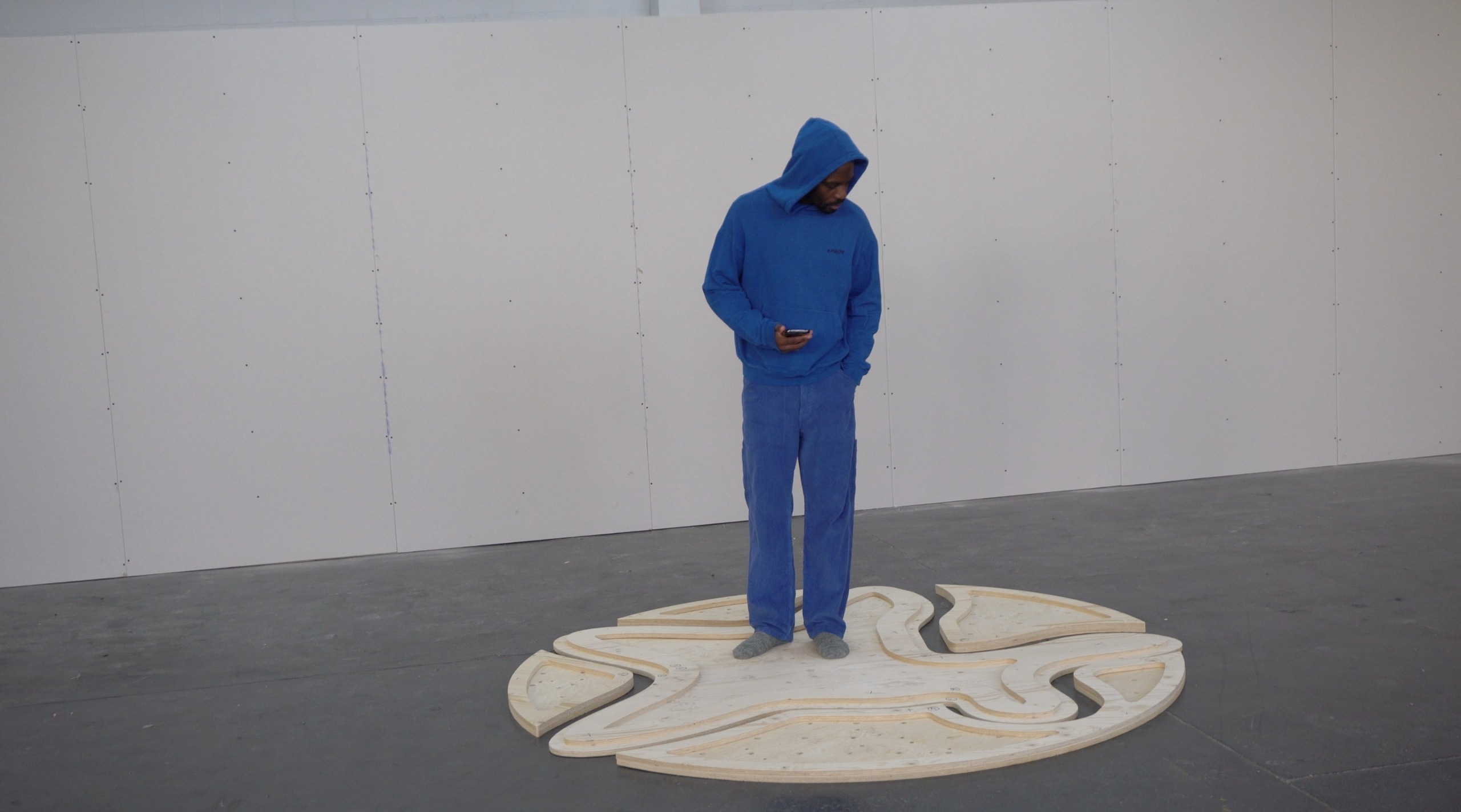 A young man poses in his industrial studio dressed in casual smartwear, sitting or standing in front of a gray wall with his sculptural creations.