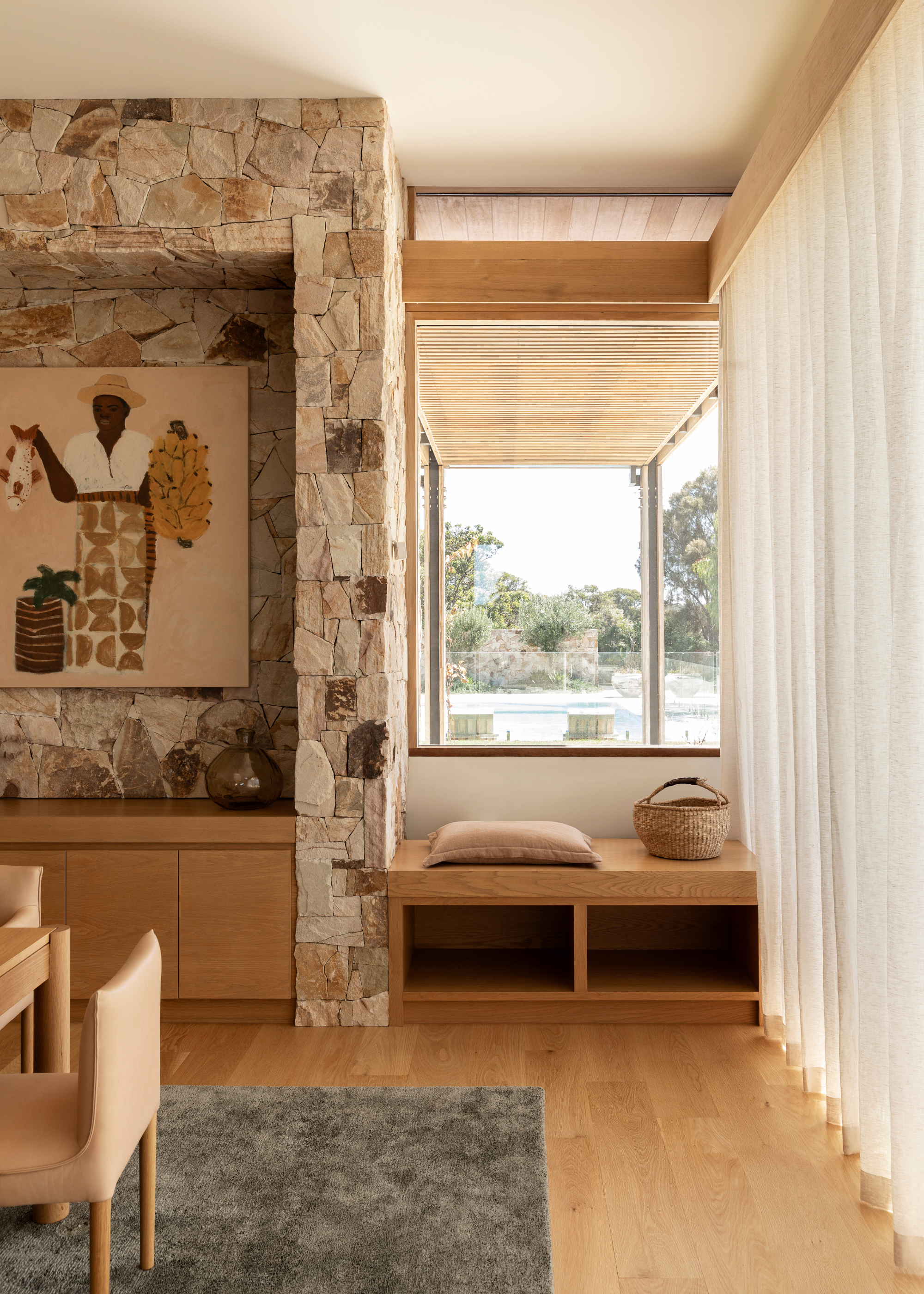 A minimalist living room with wood accents, a stone walled nook with a painting, and a beige dining set