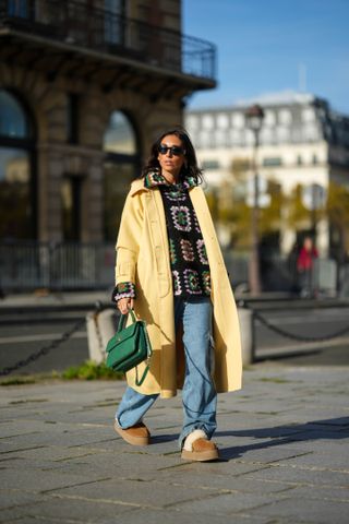 Gabriella Berdugo wears black sunglasses from Versace, gold earrings, a black / brown / green / purple / white print pattern puffy wool / zipper sweater from Gestuz, a pale yellow long coat from American vintage, a green shiny leather Coco Handle handbag from Chanel, silver rings, blue denim cargo large pants, camel suede with white sheep interior platform mules from UGGs, during a street style fashion photo session, on November 02, 2022 in Paris, France