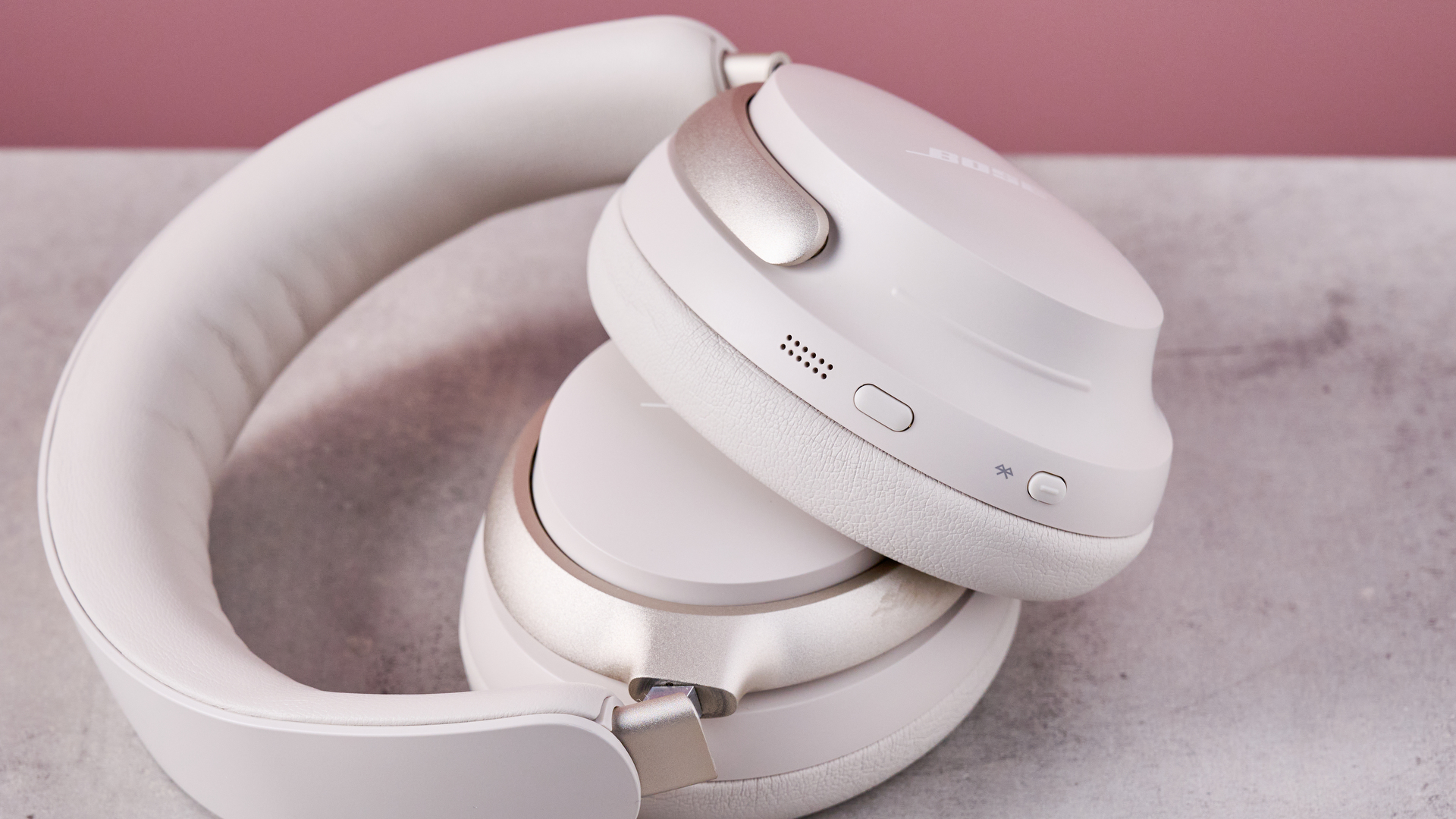 Bose QuietComfort Ultra Headphones on a gray surface with a pink background, showing the controls on the earcup